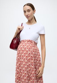 Haut blanc à manches courtes côtelées associé à une jupe longue à motifs rouges et crème. Le sujet tient un petit sac à main rouge. Texture lisse.