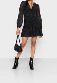 Robe mini noire avec de longues manches bouffantes transparentes, un corsage ajusté et un ourlet à volants. Associée à des bottines noires et à un petit sac.