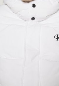 Giacca imbottita bianca con collo alto, dotata di bottoni neri e un piccolo logo. Texture liscia con sezioni trapuntate per l'isolamento.