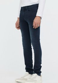 Jean slim bleu foncé en denim extensible, avec un design classique à cinq poches, associés à des baskets blanches et grises.