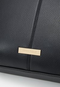 Surface en cuir noir texturé avec une couture verticale et une petite plaque rectangulaire dorée gravée du nom de la marque Anna Field.