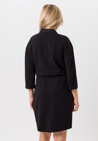 Zwarte jurk tot de knie met driekwartmouwen en een elastische taille. Beschikt over een klassieke kraag en opgerolde mouwmanchetten. Gladde textuur.