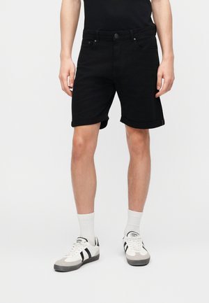 Homme portant un short noir à ourlet retroussé, associé à des chaussettes blanches montantes et des baskets blanches à rayures noires, debout devant un fond uni.