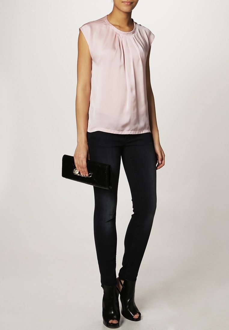 Femme portant un chemisier sans manches rose pâle, un jean skinny noir, des talons ouverts noirs, et tenant une pochette noire.