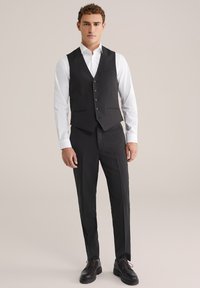 Melnā pielāgotā veste ar piecām pogām, valkāta virs baltas krekliņas. Apvienota ar melniem pieguļošiem biksēm un spīdīgiem melniem apaviem. Vienkāršs dizains.