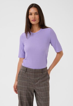 Femme aux longs cheveux sombres portant un haut violet clair à manches courtes et un pantalon marron à carreaux taille haute, debout devant un fond uni.