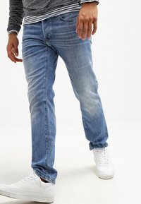 Person i ljusblå slitna jeans, vita sneakers, en grå långärmad tröja och en randig skjorta under som står på ett vitt golv.