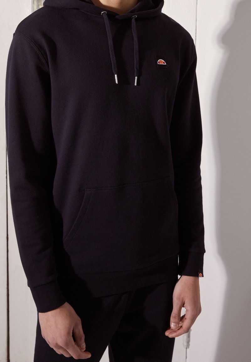 Personne portant un sweat à capuche noir avec poche avant et cordons, petit logo en demi-cercle orange et rouge sur la poitrine et la manchette du poignet.