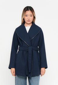 Femme se tenant devant un fond blanc, portant un manteau bleu marine ceinturé sur un haut blanc et un jean bleu clair.