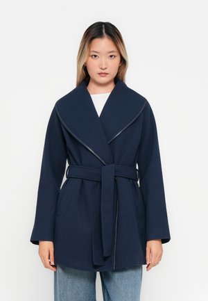 Femme se tenant devant un fond blanc, portant un manteau bleu marine ceinturé sur un haut blanc et un jean bleu clair.