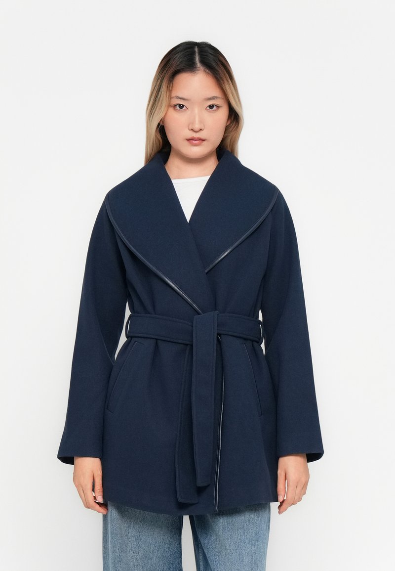 Femme se tenant devant un fond blanc, portant un manteau bleu marine ceinturé sur un haut blanc et un jean bleu clair.