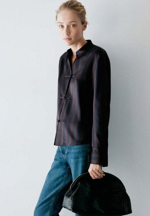 Femme aux cheveux clairs, portant un chemisier noir rayé et un jean bleu, tenant un sac à main noir texturé sur un fond clair uni.