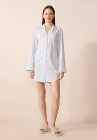 Abito camicia a righe azzurre e bianche con patta di bottoni, maniche lunghe e orlo arrotondato; abbinato a ciabatte beige.