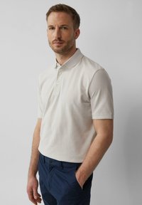 Polo shirt léger beige à manches courtes, avec un col à boutons et une finition texturée, associé à un pantalon bleu marine. Design simple et classique.