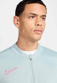 Svetlo modra zip-up jakna iz gladkega materiala, ki ima rožnati Nike logo na levem prsnem delu in visok ovratnik.