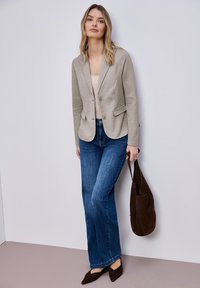 Femme debout contre un mur portant un blazer beige, un haut clair, un jean bleu, des chaussures foncées pointues, tenant un sac à main en daim brun foncé.