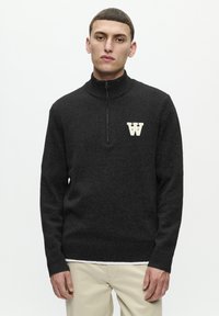Pulover negru cu fermoar, realizat dintr-un material moale, cu un guler înalt cu panglici și un logo "W" de culoare deschisă pe piept. Mânecile și tivul sunt cu panglici.