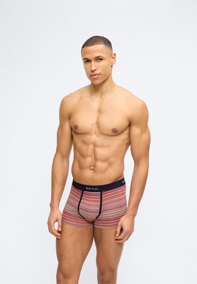 Homme musclé torse nu portant un boxer multicolore à rayures avec une ceinture noire et un logo, debout devant un fond clair uni.