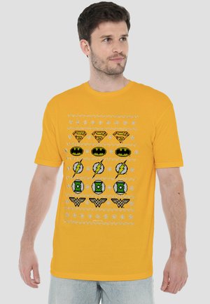 Mann trägt ein gelbes T-Shirt mit gemusterten Reihen von Superhelden-Logos, darunter Superman, Batman, Flash, Green Lantern und Wonder Woman.