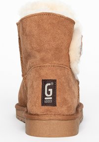 Bruine suède enkellaars met een fluffy witte voering, textuur op de zijkant met stiksels en een rechthoekig zwart logo-opdruk op de achterkant.