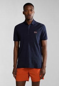 Polo shirt blu navy con una patta a tre bottoni e un piccolo emblema della bandiera norvegese sul petto, abbinato a pantaloni corti arancioni vivaci.