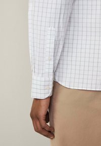 Camicia bianca a maniche lunghe con motivo a griglia grigia, polsini con bottone e orlo dritto. Il tessuto appare liscio e leggero.