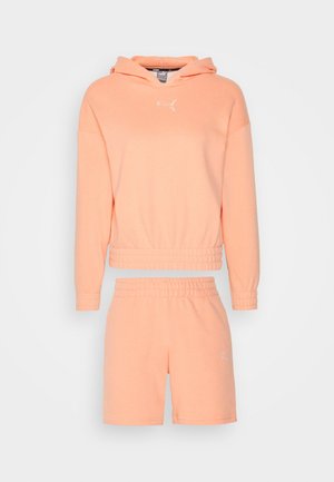 Sæt med lyserød Puma hættetrøje og elastiske taljeshorts med hvide Puma-logoer på bryst og lår, vist på en ensfarvet baggrund.