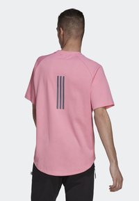 Růžové tričko s krátkými rukávy a kulatým výstřihem, na zádech výrazné tmavě šedé logo se třemi pruhy. Měkká, hladká textura.