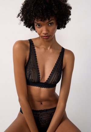 Un bralette en dentelle noire et un culotte taille haute assortie présentent des bords festonnés, des motifs floraux et une texture douce, conçus avec un décolleté en V profond.