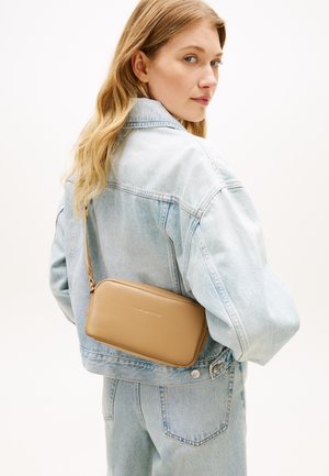 Νεαρή γυναίκα που φοράει ανοιχτό μπλε τζιν μπουφάν και τζιν παντελόνι, κρατώντας μια μπεζ τσάντα Tommy Hilfiger crossbody στην πλάτη της.