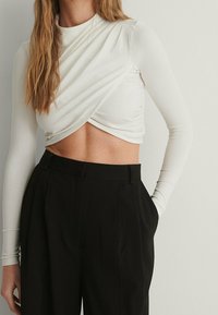 Weißes langärmliges Crop-Top mit einem verdrehten Frontdesign, kombiniert mit hochtaillierten schwarzen Hosen mit Falten und einer Seitentasche.