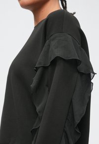 Zwarte trui met een ronde hals en ruches van doorschijnende stof op de mouwen, die een gelaagde textuur en een elegante silhouet creëren.