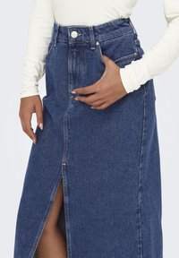 Jeanskjol med hög midja, framslits och knäppning. Blåtvätt med kontrasterande sömmar. Modellen har handen nära sidofickan.