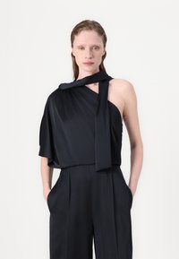 Top preto sem mangas com uma silhueta drapeada, apresentando um detalhe atado no ombro, combinado com calças pretas de cintura alta.