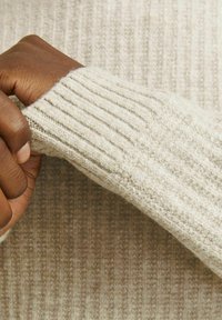 Beige bordázott pulóver ujja texturált anyaggal és hajtott mandzsettával, amely egy puha, kötött anyagot mutat be, valamint közelről látható a kéz szorítása.