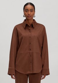 Vrouw in een bruine button-up shirt en bijpassende broek, staand tegen een effen lichtgrijze achtergrond, recht vooruit kijkend.