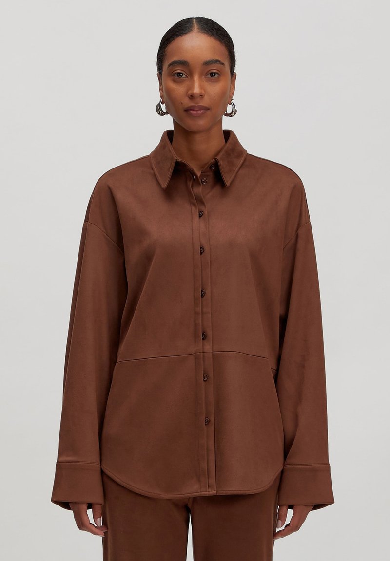Vrouw in een bruine button-up shirt en bijpassende broek, staand tegen een effen lichtgrijze achtergrond, recht vooruit kijkend.