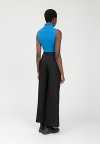 Blauwe mouwloze coltrui gecombineerd met hoge zwarte wijde broek. De broek heeft twee achterzakken en een gladde textuur.