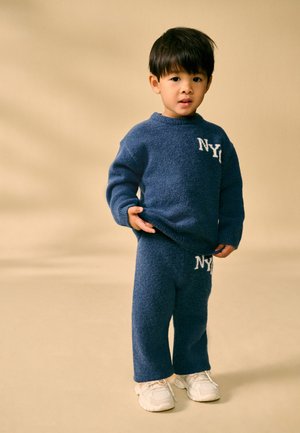 Kleines Kind trägt blauen Strickpullover und Hose mit weißen "NY"-Logos, steht vor beigem Hintergrund und hebt den Pullover leicht an.