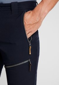 Ruka odpočívá uvnitř částečně zipované kapsy tmavě modrých kalhot, přičemž jsou vidět dva kovové zipy s hnědými poutky.