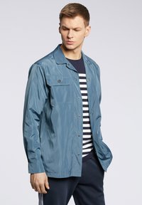 Jasnoniebieska, lekka kurtka o gładkiej fakturze, z długimi rękawami, przednimi kieszeniami i zatrzaskami, noszona na granatowej i białej koszuli w paski.