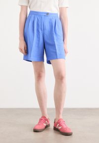 Blå linne-shorts med veckad design och knappstängning fram, i kombination med rosa sneakers med en vit detalj och grå sula.