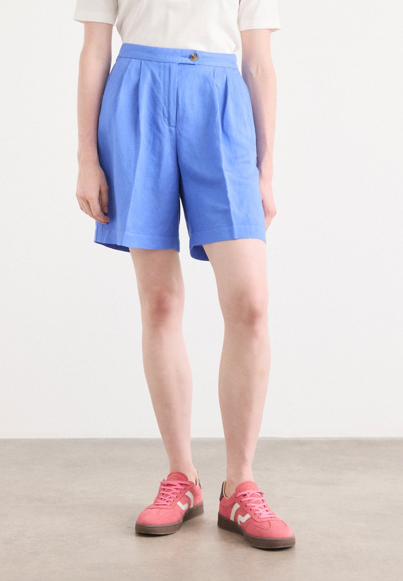 Blå linne-shorts med veckad design och knappstängning fram, i kombination med rosa sneakers med en vit detalj och grå sula.