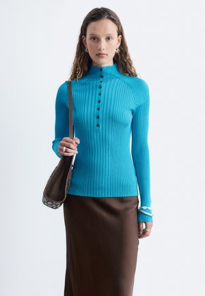 Vrouw die een blauw geribd langwerpig shirt met knopen draagt, een bruine rok, zilveren oorringen, armbanden en een donkerbruine handtas bij zich heeft.