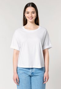 Een glimlachende vrouw met lang haar draagt een losse witte t-shirt en lichtblauwe jeans, staand tegen een neutrale achtergrond.