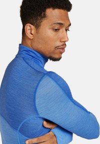 Top deportivo azul con cuello alto, mangas largas y una tela ligera texturizada con tonos de azul contrastantes en un diseño de panel.