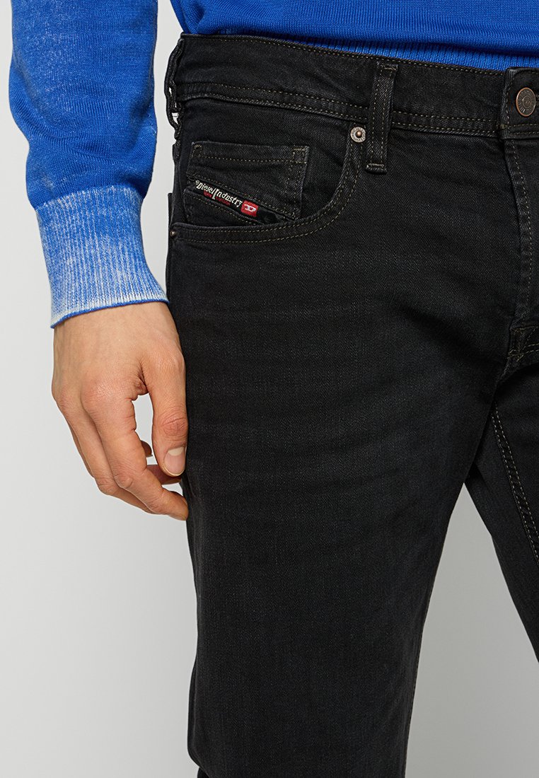 Jeans en denim noir avec un design classique à cinq poches, des coutures robustes et une étiquette rouge contrastante sur la poche avant.