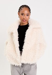 Veste en fausse fourrure de couleur crème douce, avec un large col, coupe ample et tissu texturé. Associée à un haut noir et un jean gris.