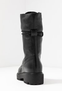 Botte noire en cuir, montant jusqu'au genou, avec une finition texturée, dotée d'un détail en nœud à l'arrière, d'une semelle en caoutchouc robuste et d'un bout arrondi.