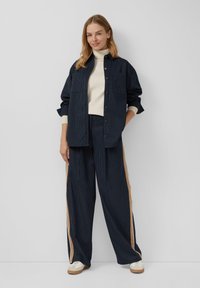 Giacca a righe blu navy sopra un dolcevita beige chiaro, abbinata a pantaloni a gamba larga con strisce sui lati color crema e marrone chiaro. Sneakers bianche.